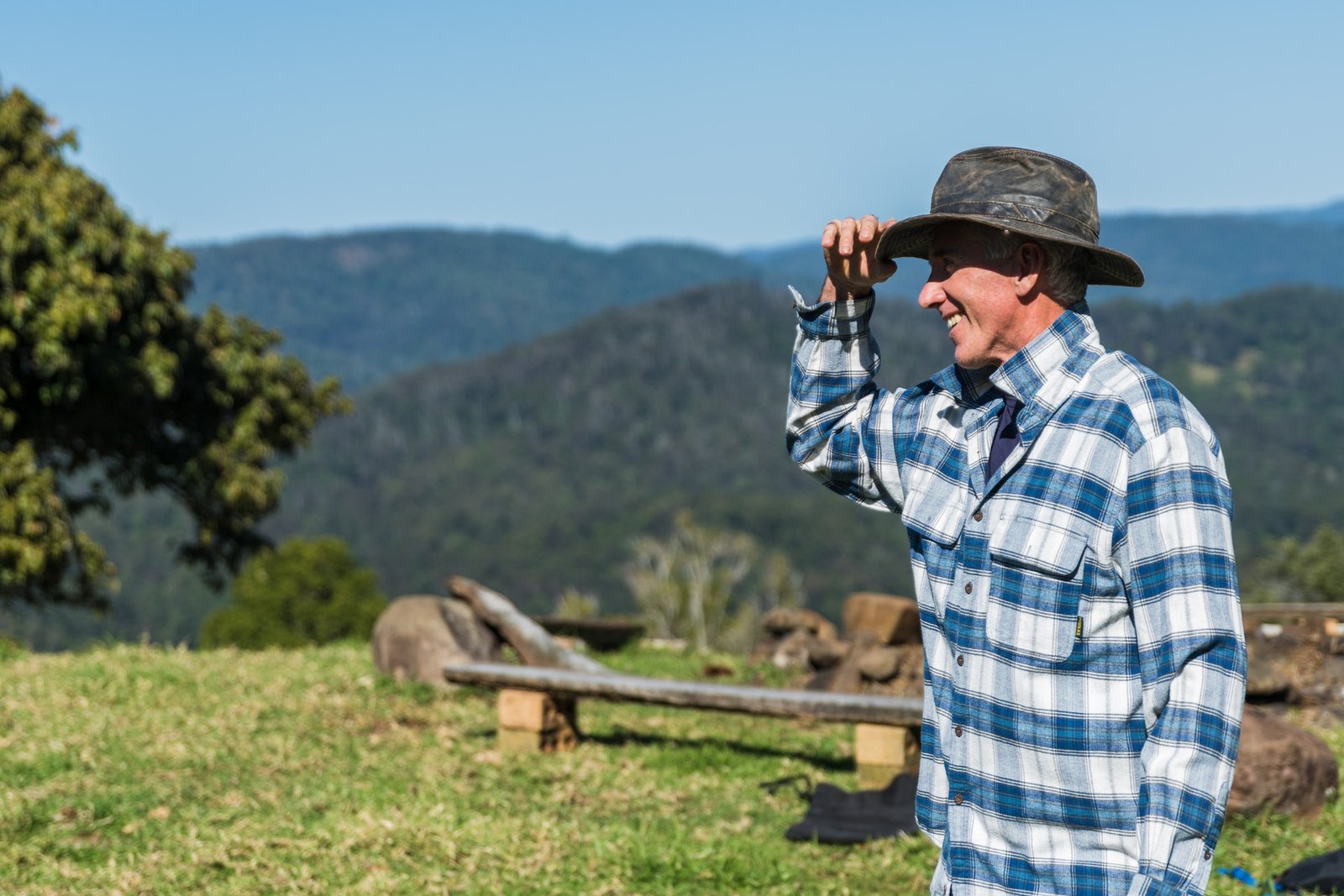 Un hombre sonriente vestido con una camisa de cuadros azules y un sombrero de ala ancha, de pie en un campo con colinas boscosas al fondo y un cielo despejado.